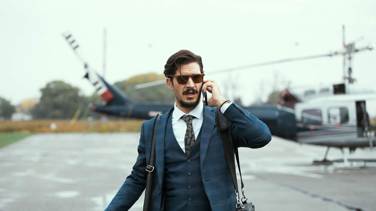 A man in a blue suit and glasses talks on a phone. A helicopter can be seen in the background.