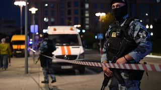 A Russian police officer in blue khakis, and a black vest and face mask holding a rifle at night. He is standing behind police tape on a street, a van and another officer are behind him.