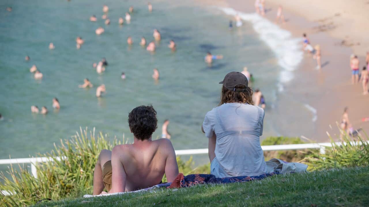 A shirtless man and a woman in a white shirt sit on a grass hill overlooking a beach