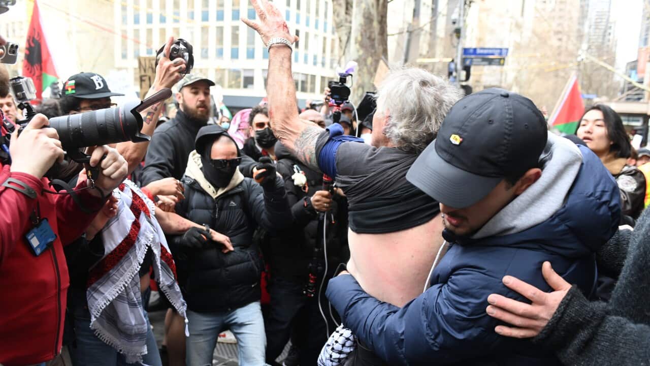 A tense street scene shows a man in a black shirt and baseball cap comforting or restraining an older man with his arm raised, surrounded by a crowd of people, some holding cameras.