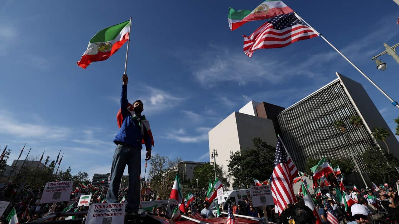 A man holding an Iranian flag high above his head stands above a crowd holding a mixture of Iranian and American flags