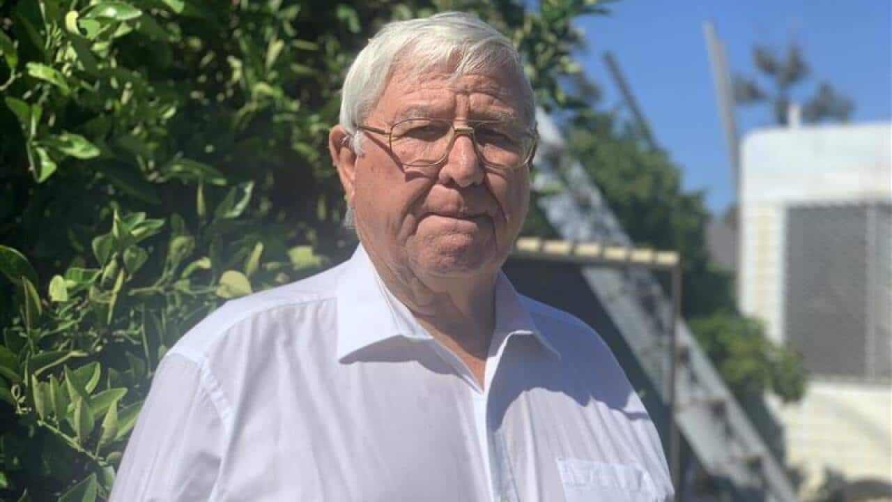 A man in a white shirt stands outside in a garden with a serious expression on his face.