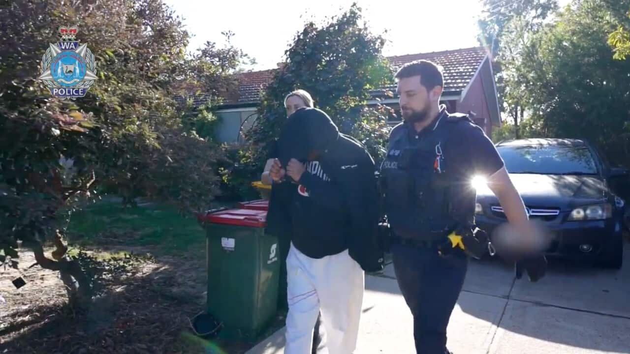 A man with his face covered by a hood is led down a suburban driveway by two police officers.