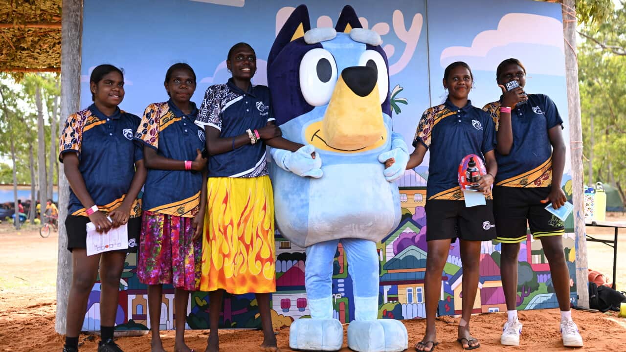 A group of young women in school shirts stand next to a person wearing a costume of the cartoon character Bluey.