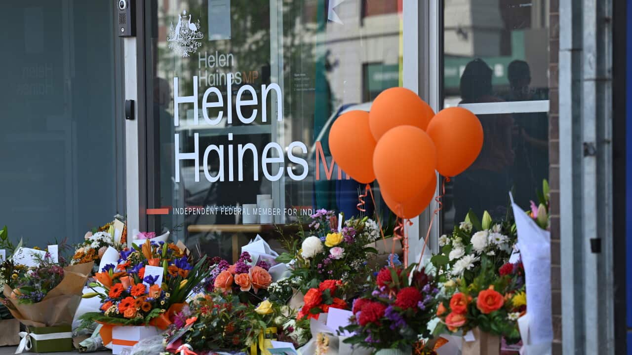 A large pile of flowers with a bunch of orange balloons sits against a window with a sticker reading "Helen Haines MP".