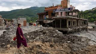 A woman standing on a muddy embankment. A building that has been partially destroyed by flood waters is behind her.