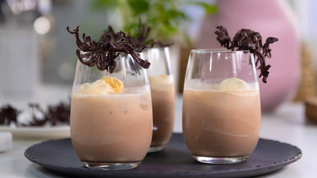 Three glasses sit on a round dark tray. The glasses are three-quarters filled with a chocolate drink, and each glass is garnished with a chocolate twirl.