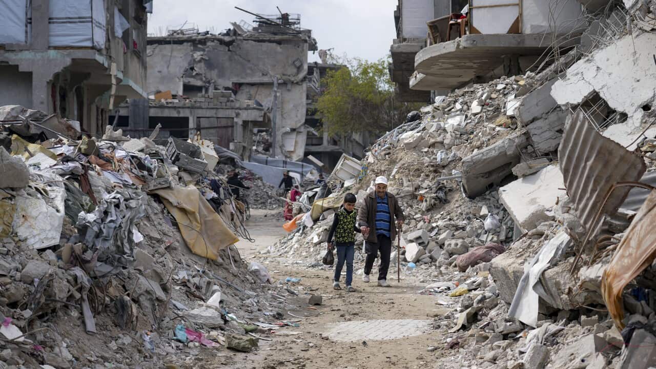 A child and an old man walk along a makeshift road in Gaza. On either side of them are large piles of rubble. Behind them are damaged buildings.