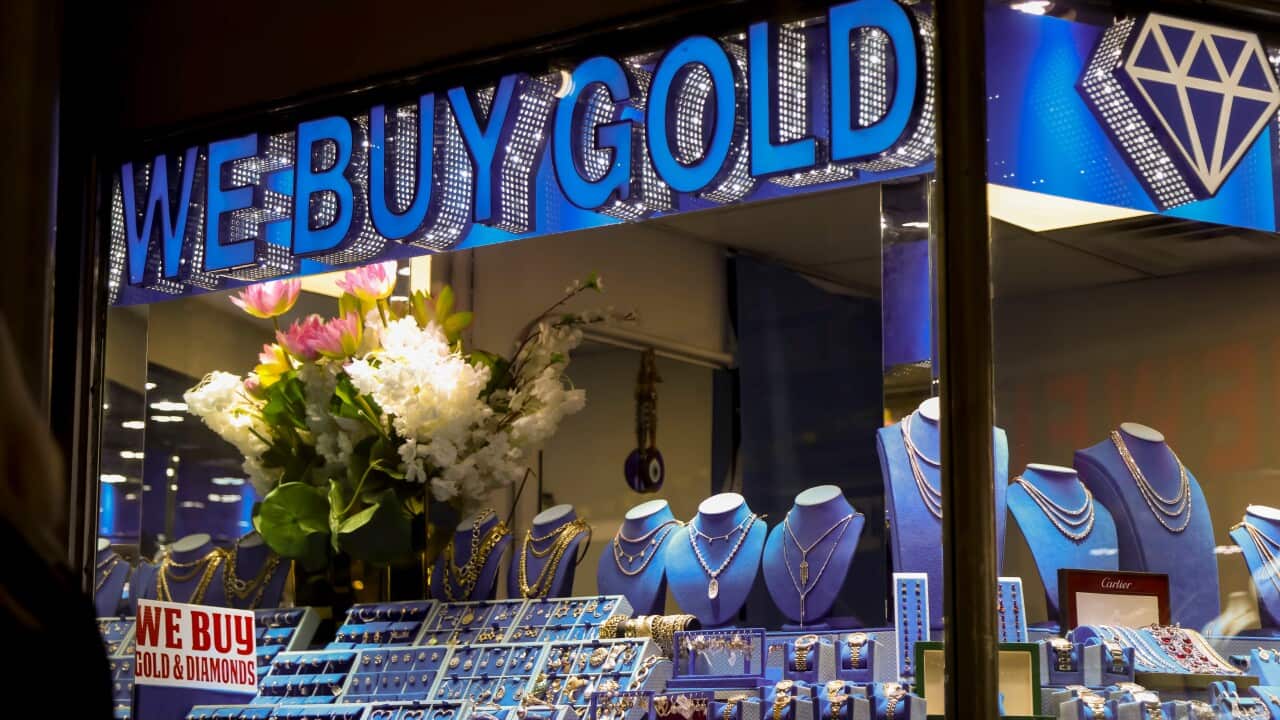 A night-time view of a jewellery store window features a large, bright blue neon sign that reads "WE BUY GOLD" and a brightly lit display of necklaces, rings, and bracelets on blue velvet busts, with a prominent floral arrangement to the left.
