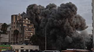 An apartment building in a city stands in the foreground, while a massive, dense plume of dark grey smoke billows into the sky directly behind it. The smoke is voluminous, suggesting a recent large-scale explosion or missile strike. In the lower foreground, a parking lot is filled with cars, and a few onlookers can be seen holding up their smartphones to record the scene. The sky is overcast and dim, adding to the somber and chaotic atmosphere of the event.