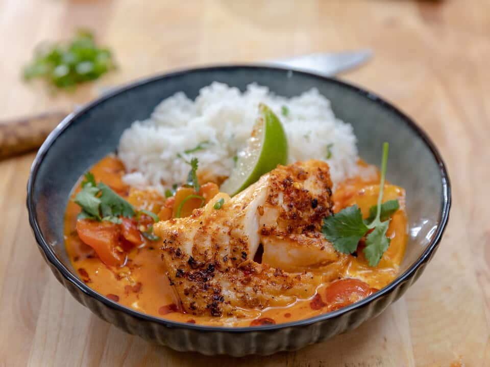A ineresting pottery bowl holds a soupy-stew with a piece of cooked white fish in an orange-ish creamy sauce, alongside white rice. The bowl is topped with a wedge of lime.