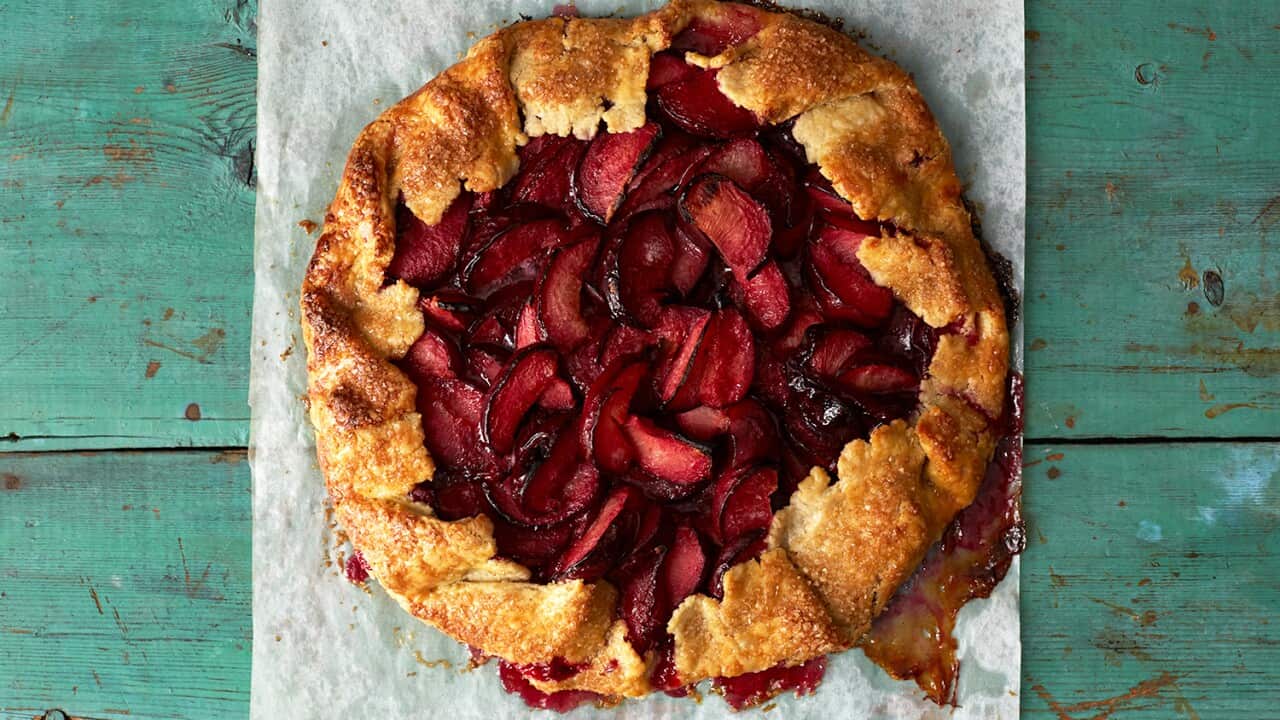 Seen from overhead, a round, rustic tart with pastry edges folded in over a fruit filling sits on baking paper, on a aqua-green wooden surface.