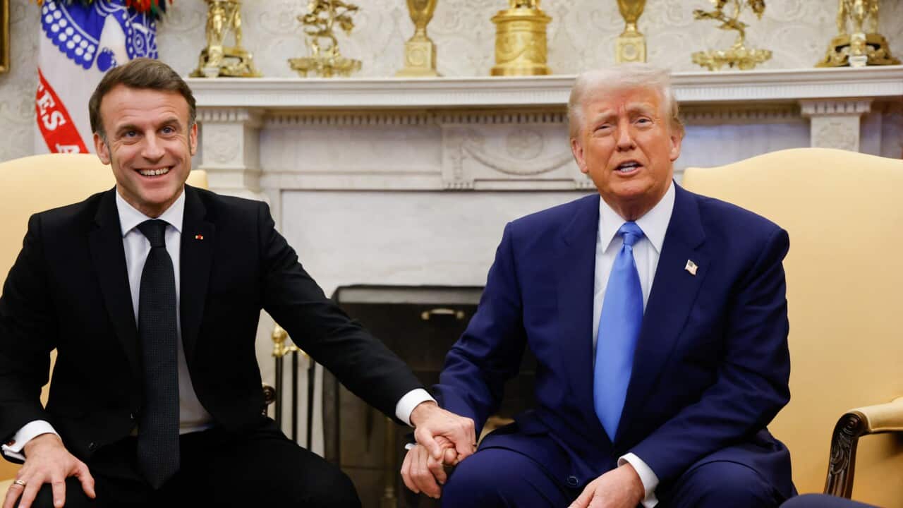 Emmanual Macron (left) wearing a black suit, holds hands with Donald Trump who is sitting next to him wearing a blue suit