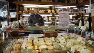 Shop interior with glass display cases filled with assorted cheeses, while a man stands behind the counter in front of shelves stocked with cheese and specialty foods.
