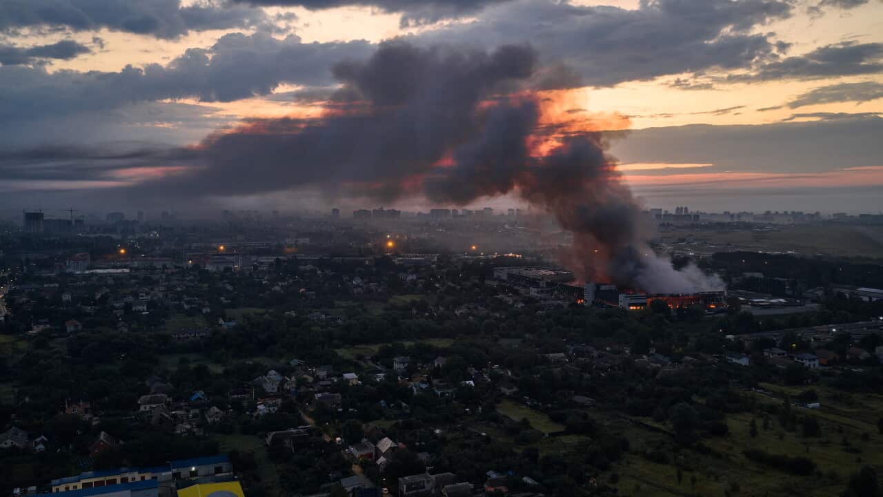 An aerial shot of a city, with smoke rising from a burning building on the right side of the image.