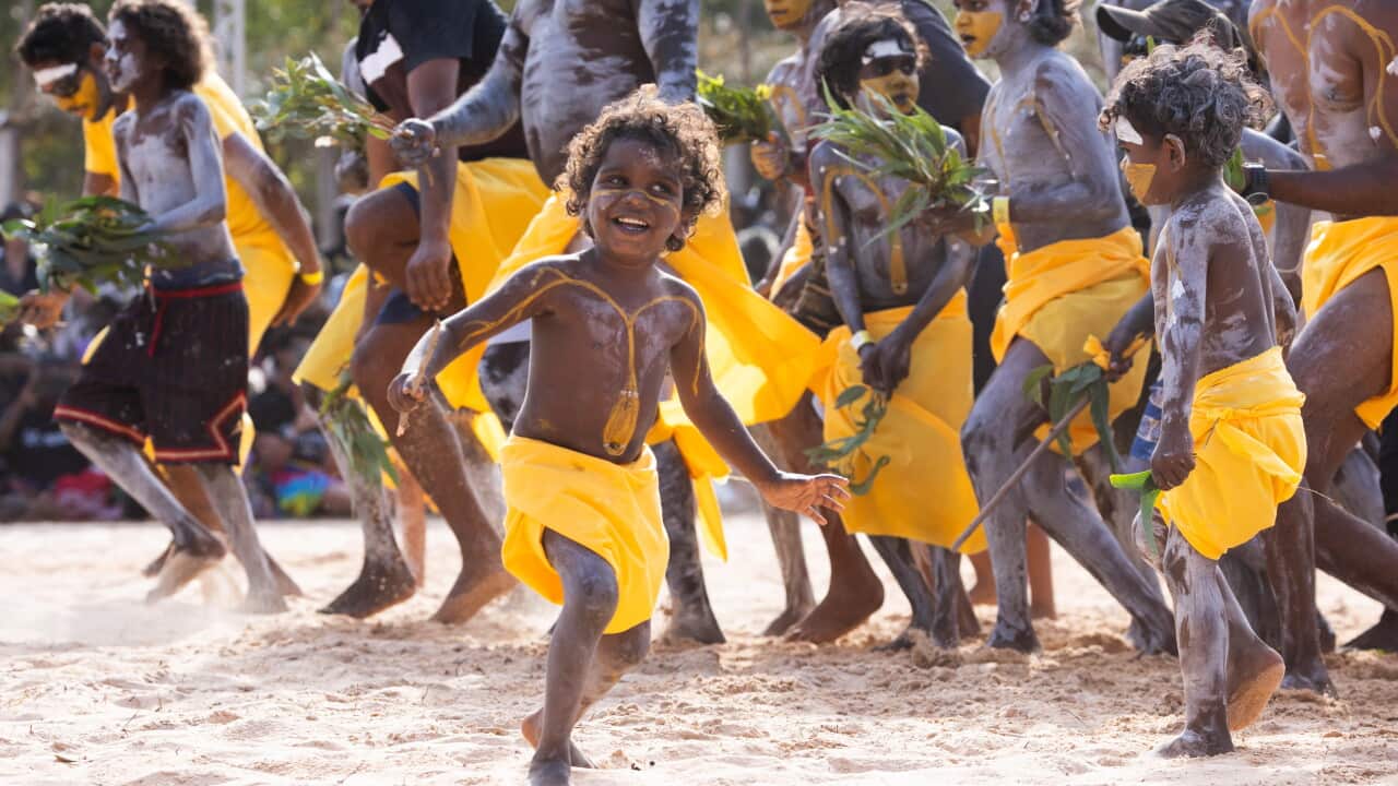 Men and young boys dressed in yellow traditional clothing and paint dancing on sand.