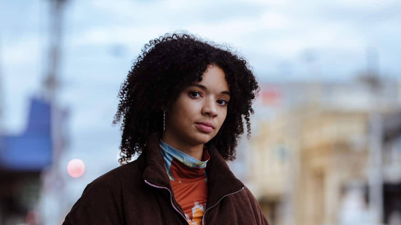A young woman with curly dark hair, wearing a heavy brown jacket, stands in front of an out-of-focus city street.
