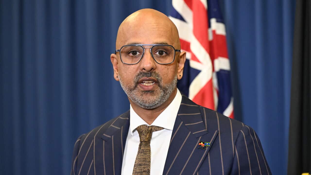A middle-aged South Asian man wearing a pin-striped suit. An Australian flag is behind him.