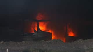 Bright orange flames blaze inside a damaged structure behind a dirt embankment beneath a thick cloud of dark smoke.