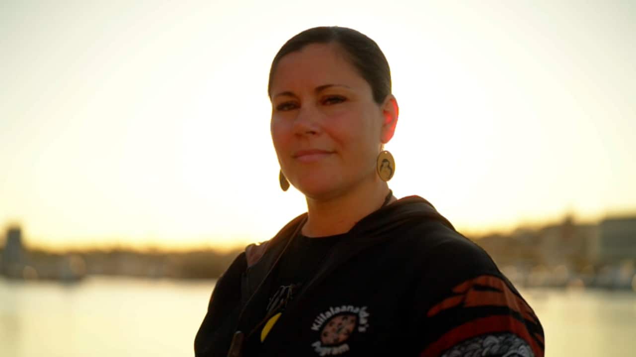 a woman with slick back hair stands mid shot smiling at the camera with the sunset in the background