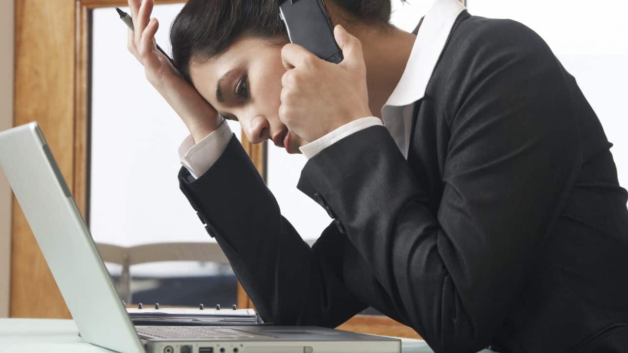 A woman in a black jacket worn over a white shirt, leaning on her hand, looking down at her computer while on the phone.