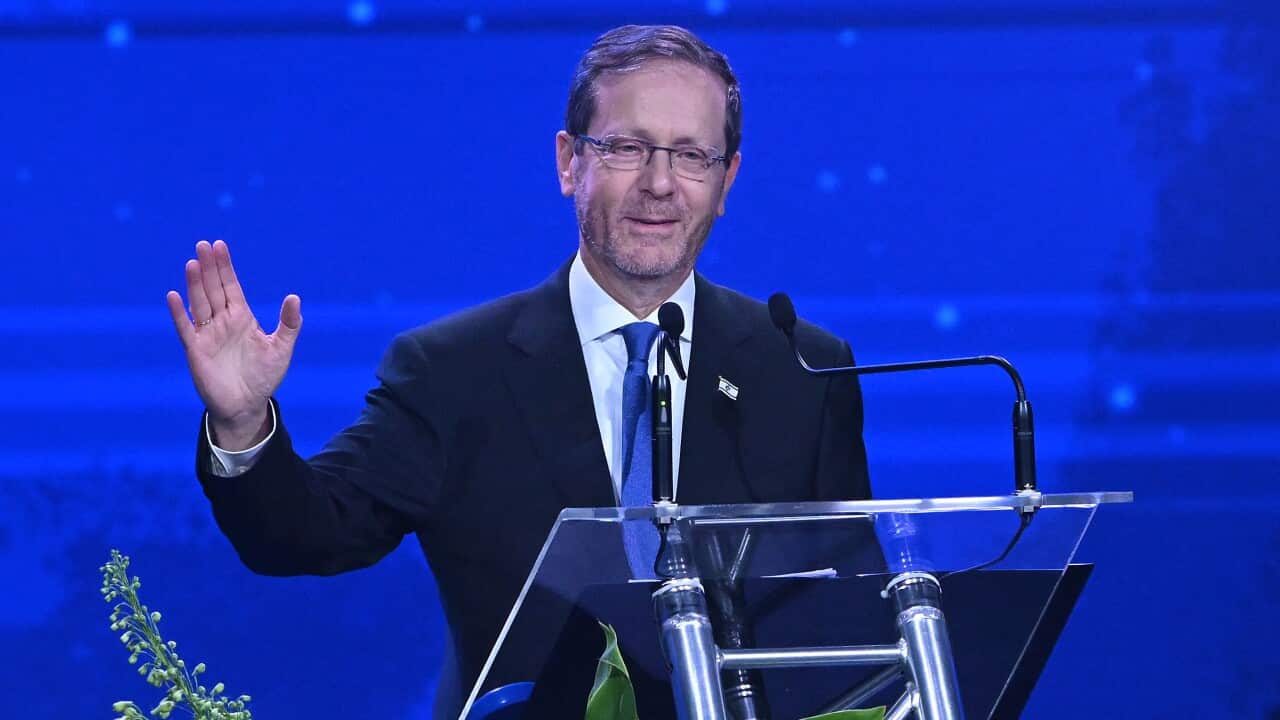 Isaac Herzog waves while speaking at a lectern against a blue background.