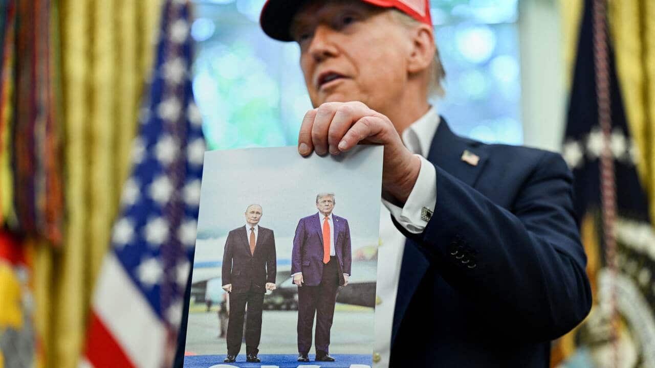 President Donald Trump, wearing a signature red MAGA cap, holds a photo of him with Russian President Vladimir Putin standing on an airport runway.