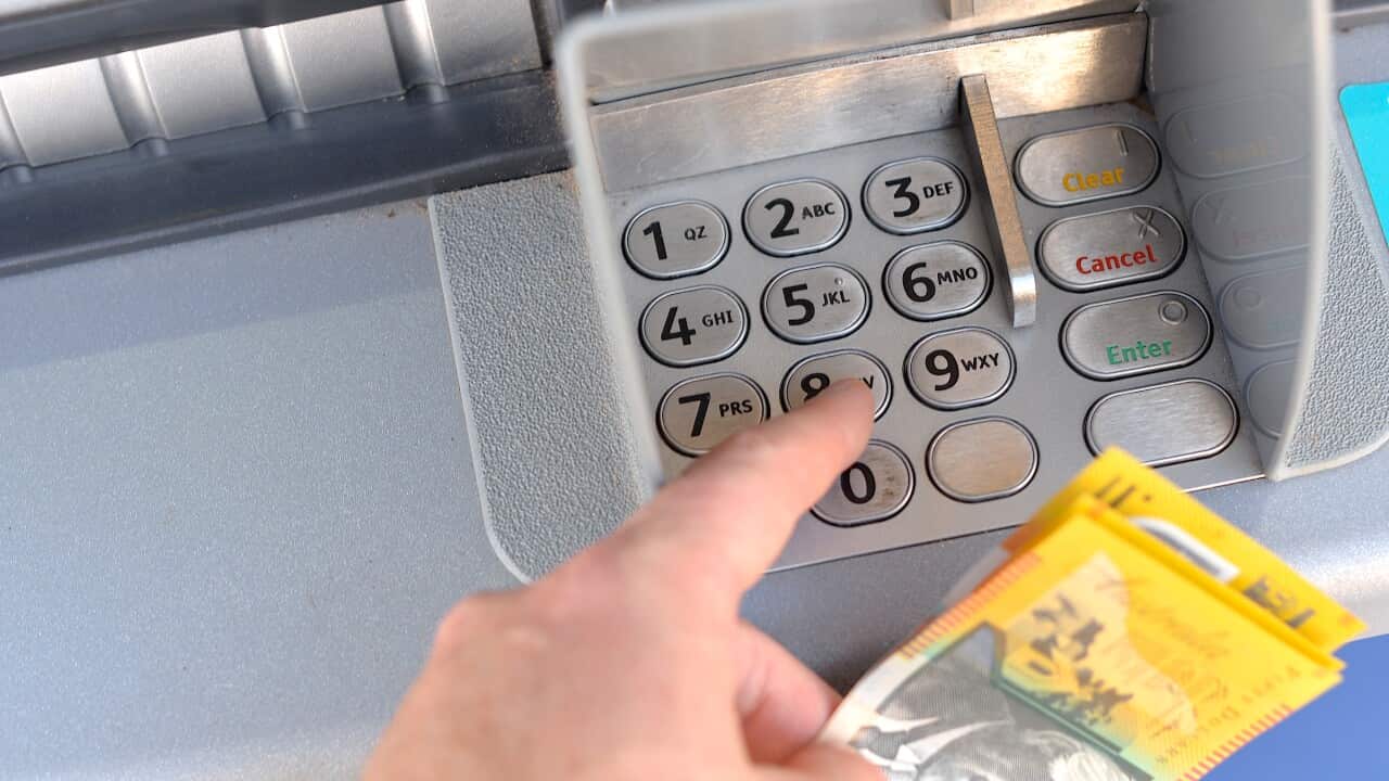 A close-up shot shows a person's finger pressing a button on an ATM keypad while they hold a fan of Australian dollar banknotes.