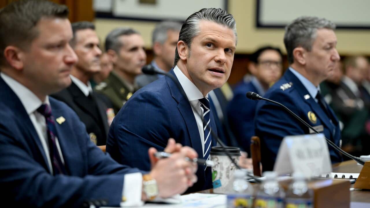 Three men in military suits sit at a bench with microphones in a crowded room.