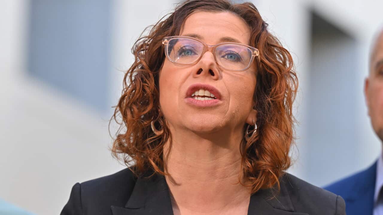 A woman with shoulder-length hair and glasses and wearing a black suit is captured as she talks to the cameras.