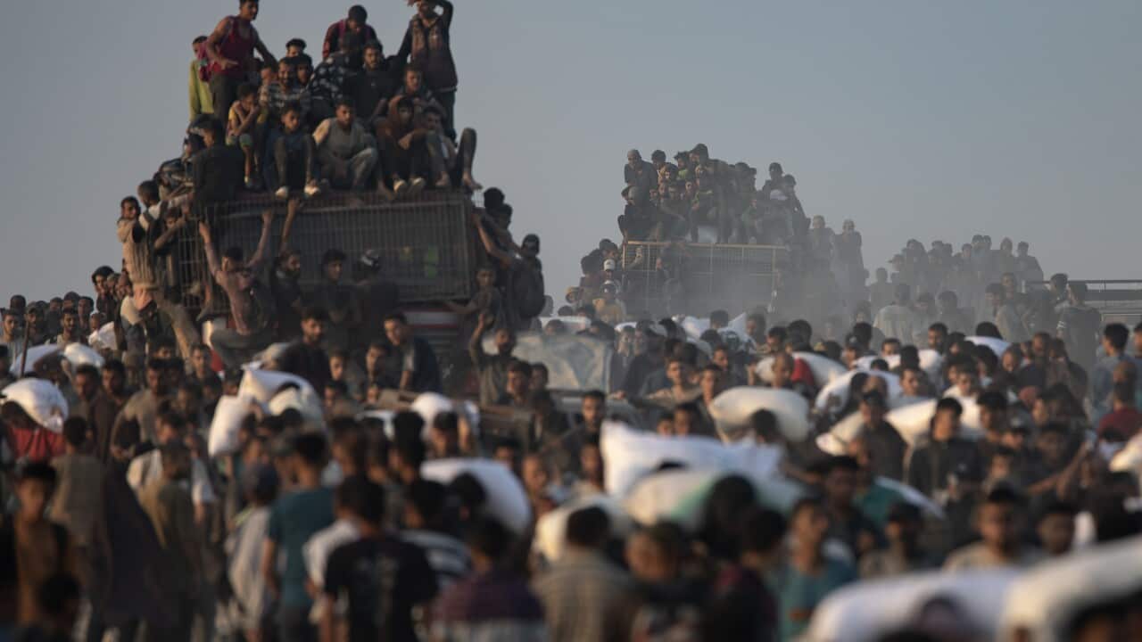 A crowded road with people carrying large white bags of flour on their heads. There are trucks overflowing with people on the same road.