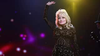 A blonde woman in a sparkling black dress raises one hand above her head.