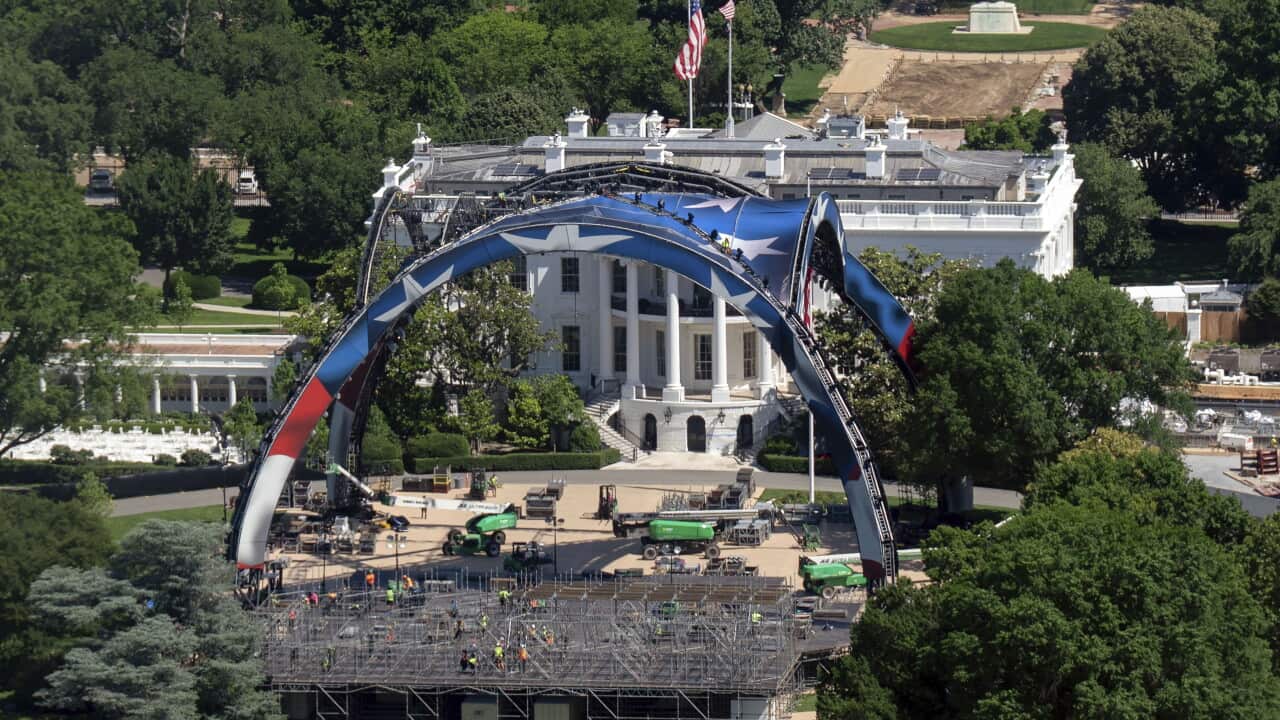 Two big red-and-blue arches atop a construction site, with a white building in the background. An US flag is flying on top of the white building and trees are seen surrounding the area