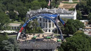 Two big red-and-blue arches atop a construction site, with a white building in the background. An US flag is flying on top of the white building and trees are seen surrounding the area