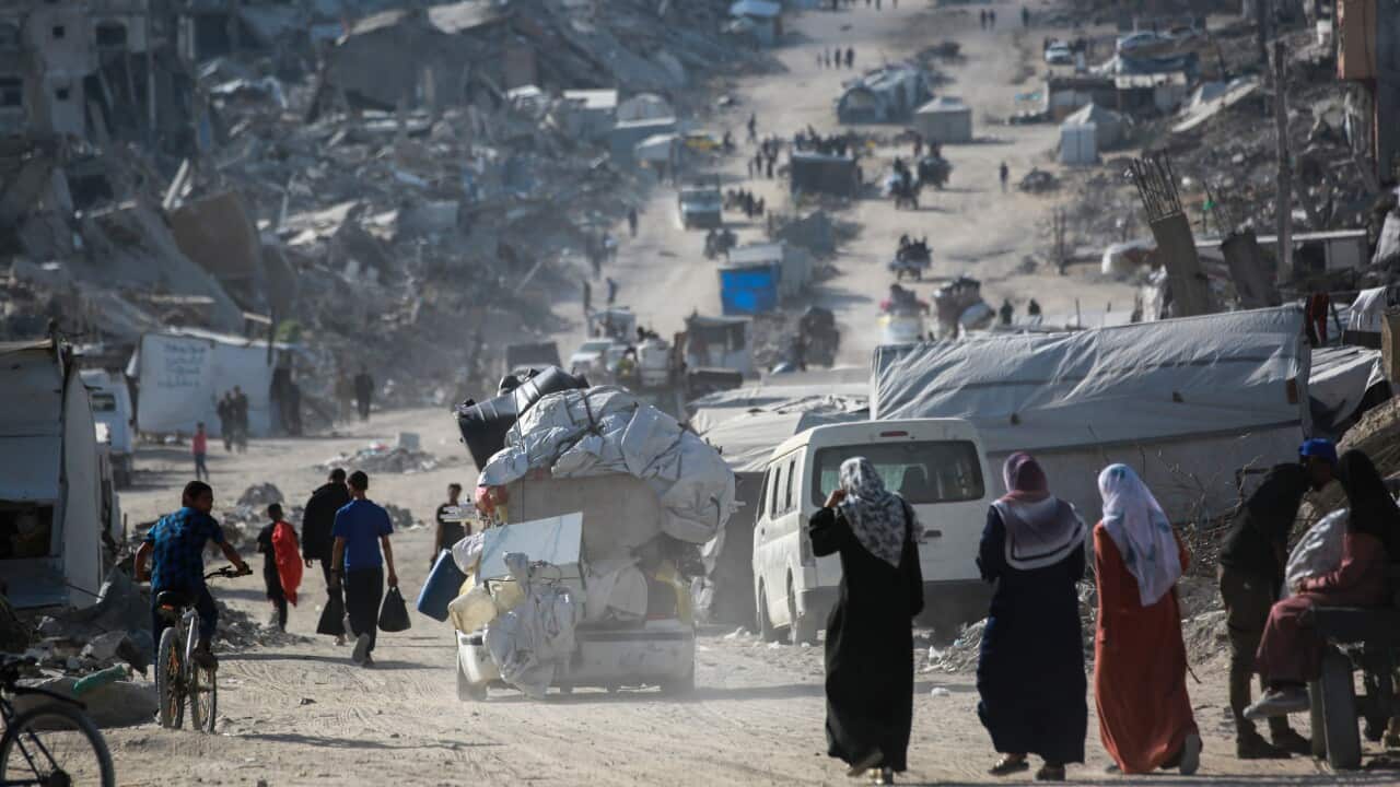 People move with their belongings along a dirt road with vehicles and destroyed buildings around them.