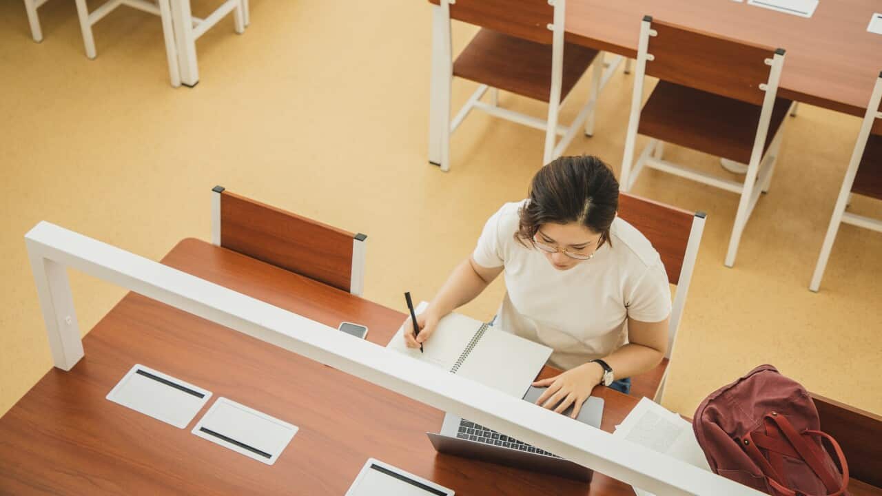 Asian woman sitting at a desk using an laptop and writing in a notebook