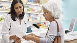 A pharmacist in a white coat reviews a prescription while consulting with an elderly woman at a pharmacy counter.