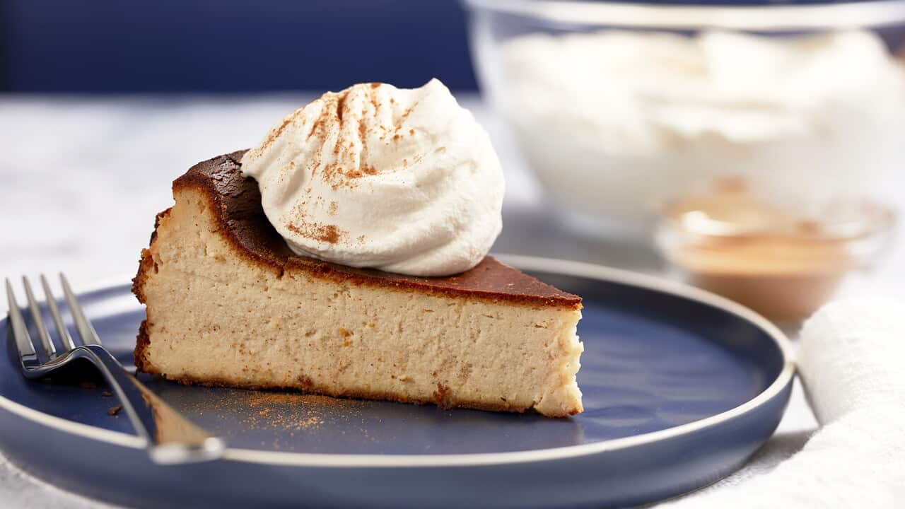 A slice of dark cream coloured cheesecake with a deep brown top sits on a wide, modern, lipped dark blue plate. A big dollop of cream sits on the cheesecake slice. A fork sits on the plate, alongside the cheesecake.