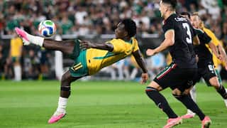 A football player in a yellow and green uniform executes a high volley kick mid-air next to an opposing player in a black jersey.