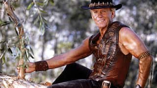 Paul Hogan as Mick Dundee in a leather vest and akubra sitting in a tree
