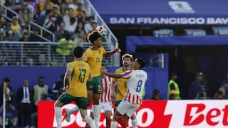 An Australian soccer player jumps high to head a soccer ball during a match against Paraguay, surrounded by teammates and opposing players in a packed stadium.