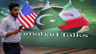 A man dressed in office attire and holding a mobile phone walks past a mural of flags from the US, Pakistan and Iran.