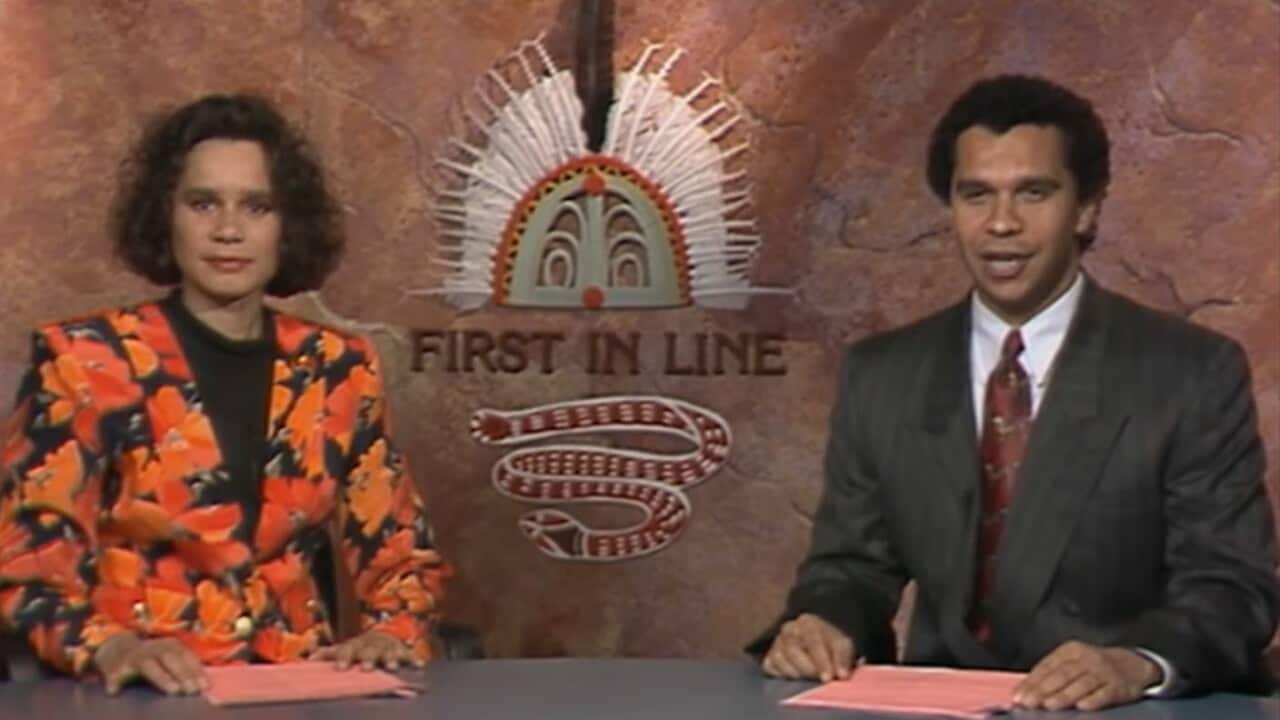 A woman in an orange and black floral jacket and a man in a dark suit with a red tie are presenting a TV program in front of a background featuring an Indigenous logo and the words "First in Line" written on it.