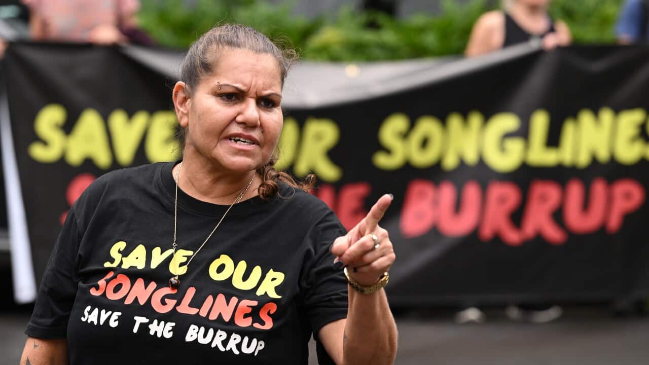A woman in a black shirt with yellow, red and white text reading: Save our songlines, save the Burrup points her finger as she stands outside.