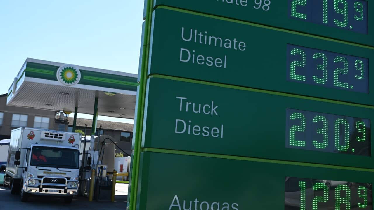 A truck filling up at a service station. There are fuel prices on a board in front.