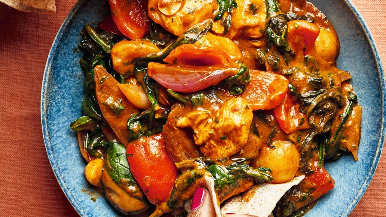 A colourful chicken and vegetable dish sits in a blue bowl. A hand can be seen scooping up some food with a folded piece of flatbread.