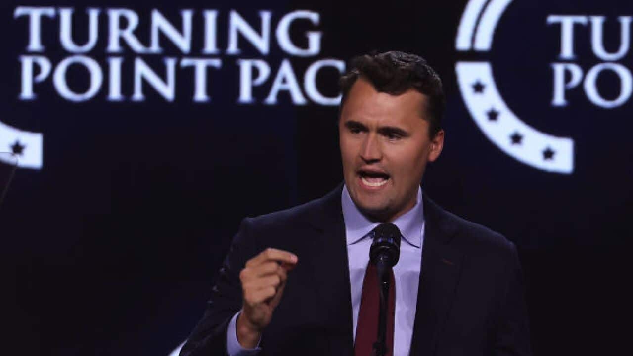 A man wearing a suit speaks to an audience in front of a dark background featuring the words "Turning Point Action" repeated across it.