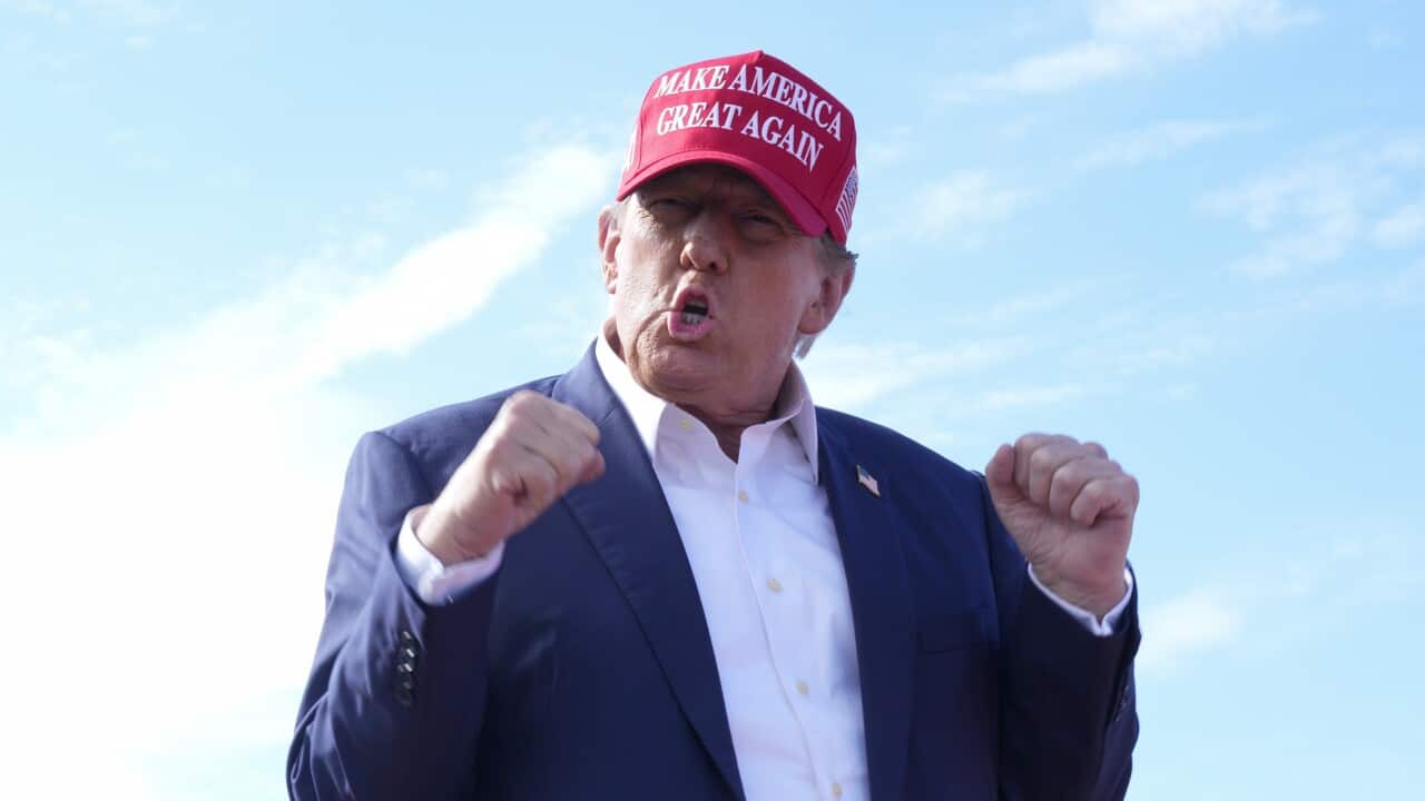 Man in red hat and blue suit pumps fists in the air. Crowd waving flags can be seen behind him.