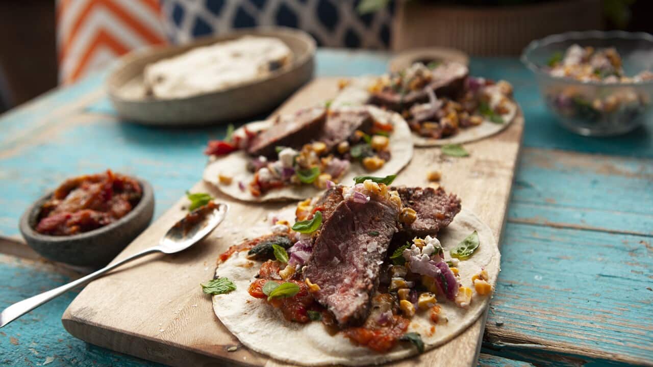 Three flatbreads sit on a wooden board, topped with sliced steak and condiments. Bowls with extra sit behind the board, all of it on an aqua blue-pained wooden tabletop.