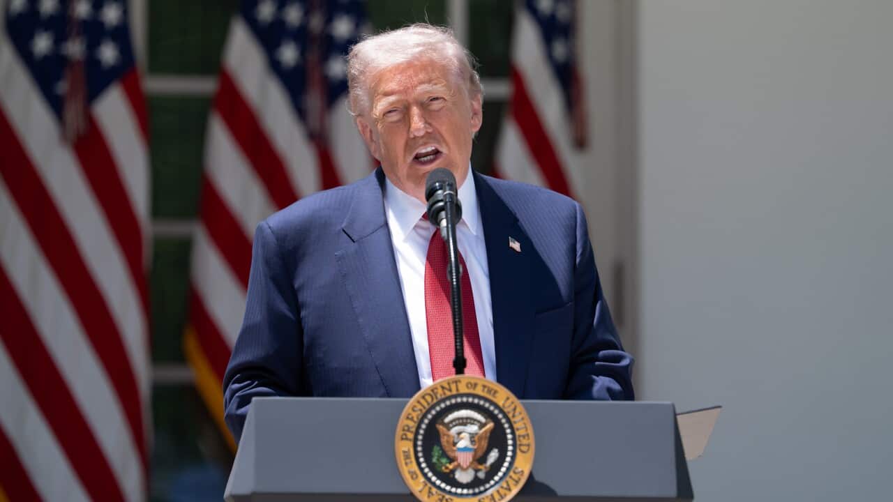 An older white man with blonde hair wearing a suit and speaking before a podium. US flags hang behind him.